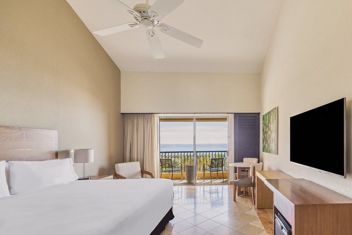A hotel room with a bed, desk, chair, TV, ceiling fan, and a balcony with a view.