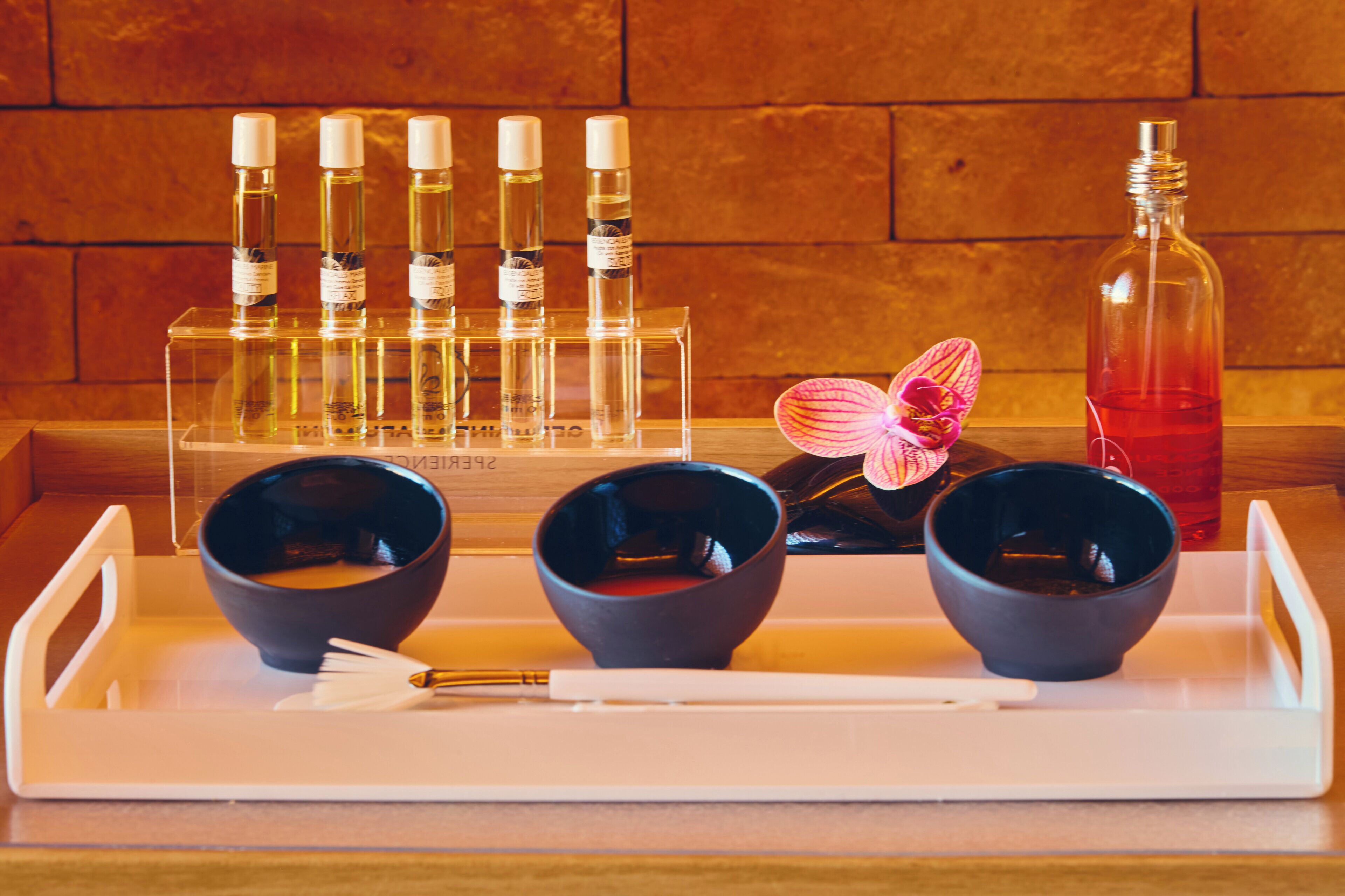 Una bandeja con tres cuencos negros, un pincel blanco y una flor rosa. Detrás de los cuencos, hay seis botellas de líquido en un recipiente transparente. A la derecha, una botella con líquido rojo.