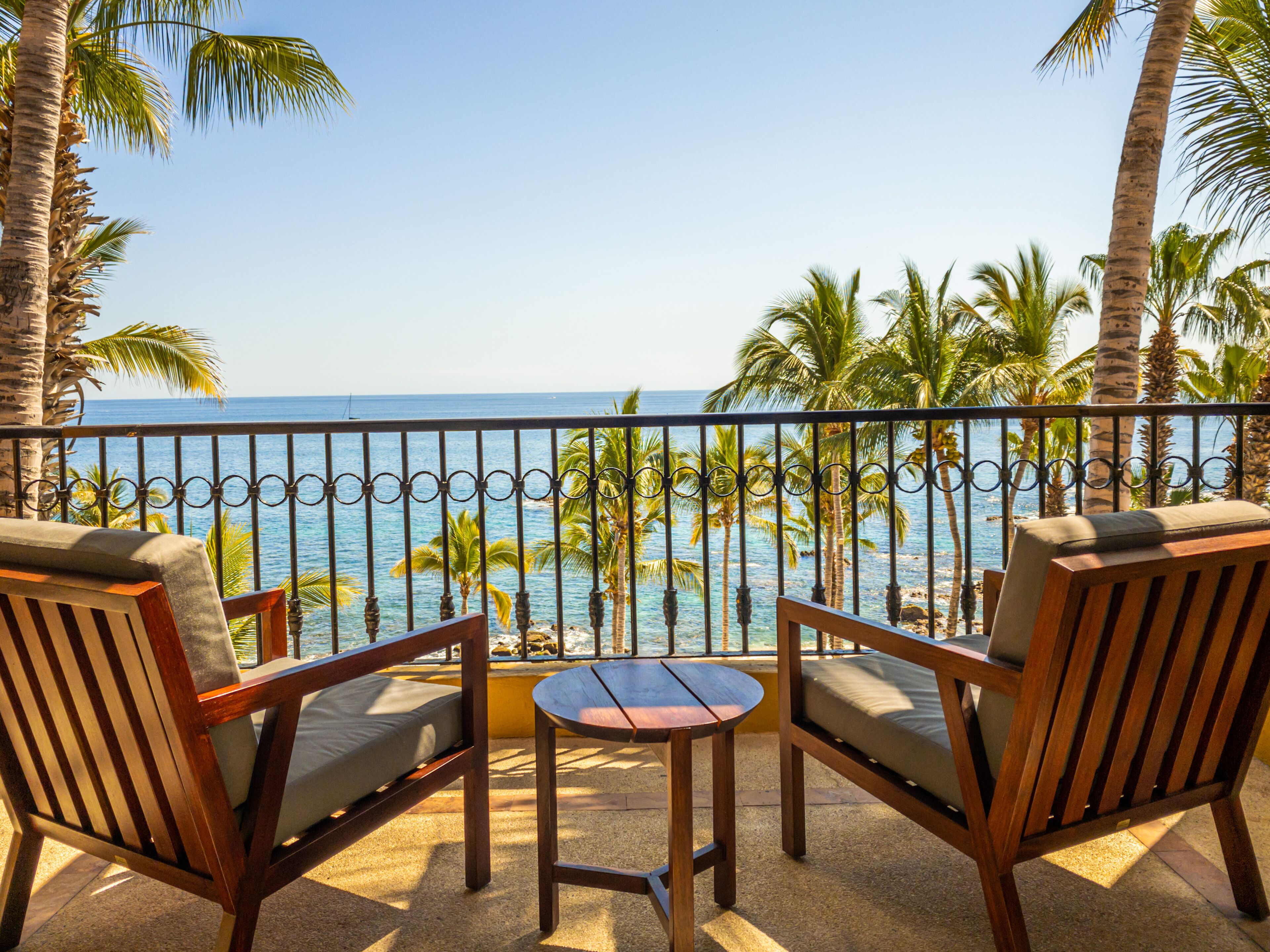 A balcony with two wooden chairs and a small table, overlooking a beach with palm trees and the ocean.