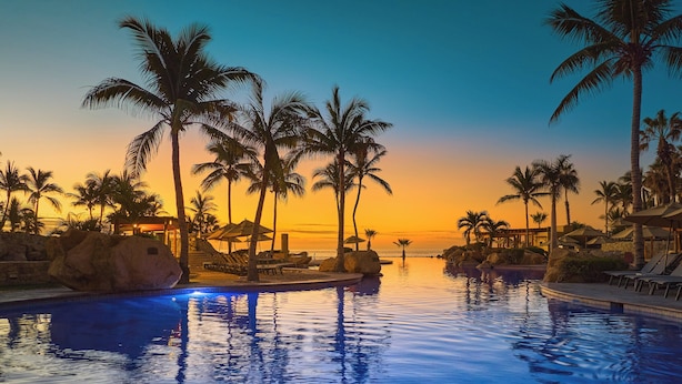 Une piscine d’un complexe hôtelier, bordée de palmiers et dotée de chaises longues, entourée de gros rochers et offrant une vue imprenable sur le coucher du soleil.