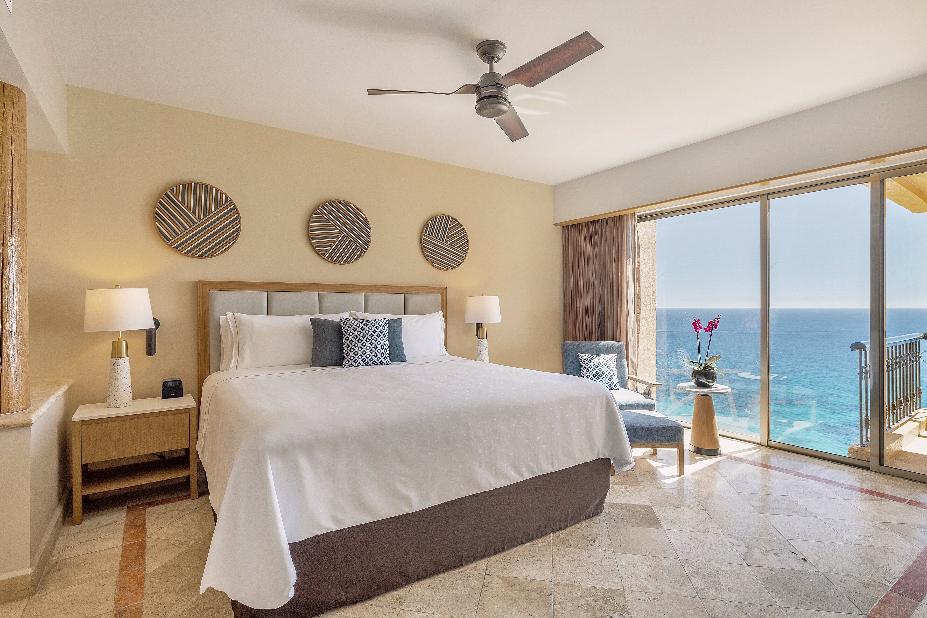 Habitación de hotel con una cama grande, dos lámparas fijadas a la pared, un ventilador de techo y vistas al océano a través de un ventanal corredero.
