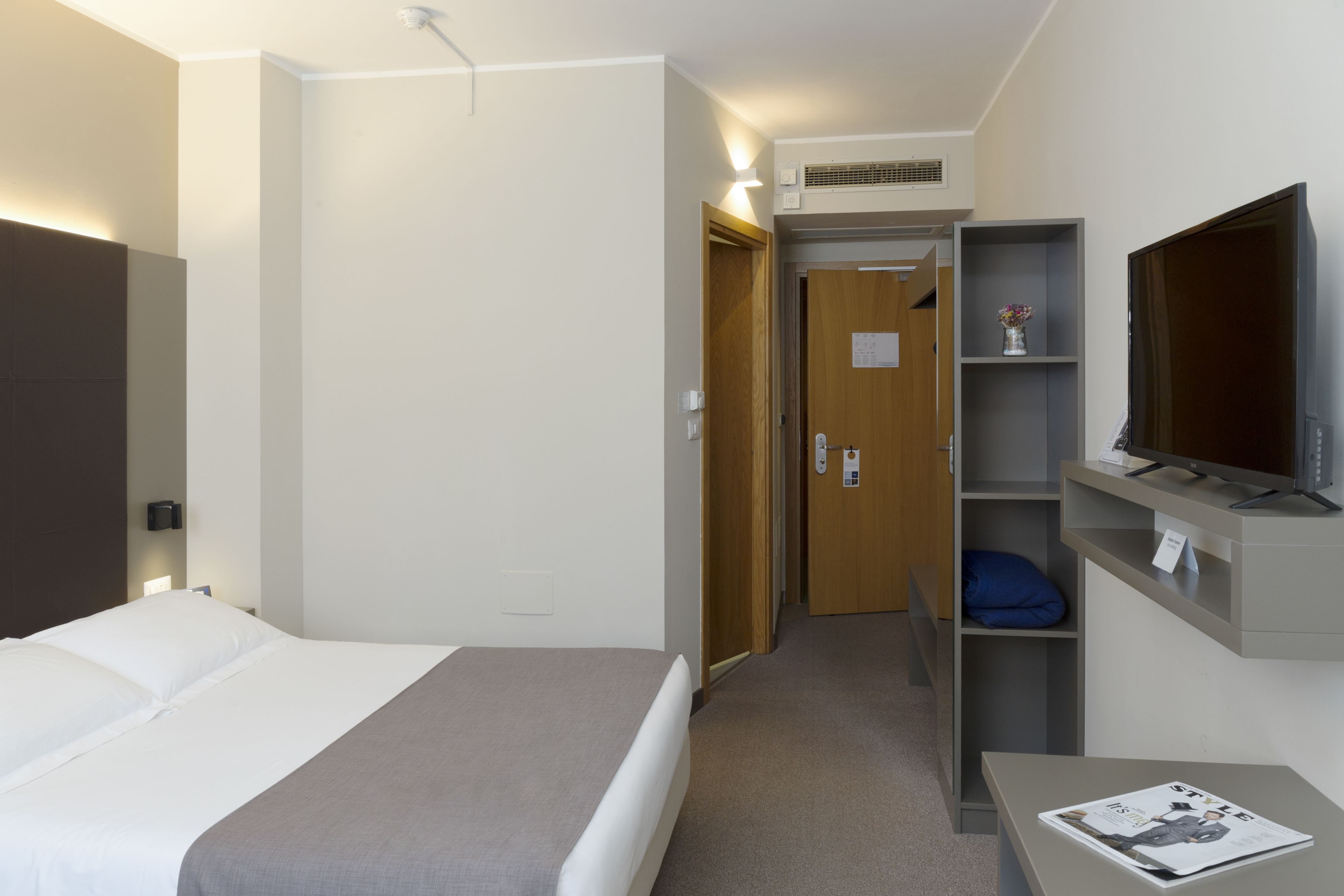 Ein Hotelzimmer mit Bett, Fernseher auf einem Schrank, Schreibtisch mit Zeitschrift und Kleiderschrank.