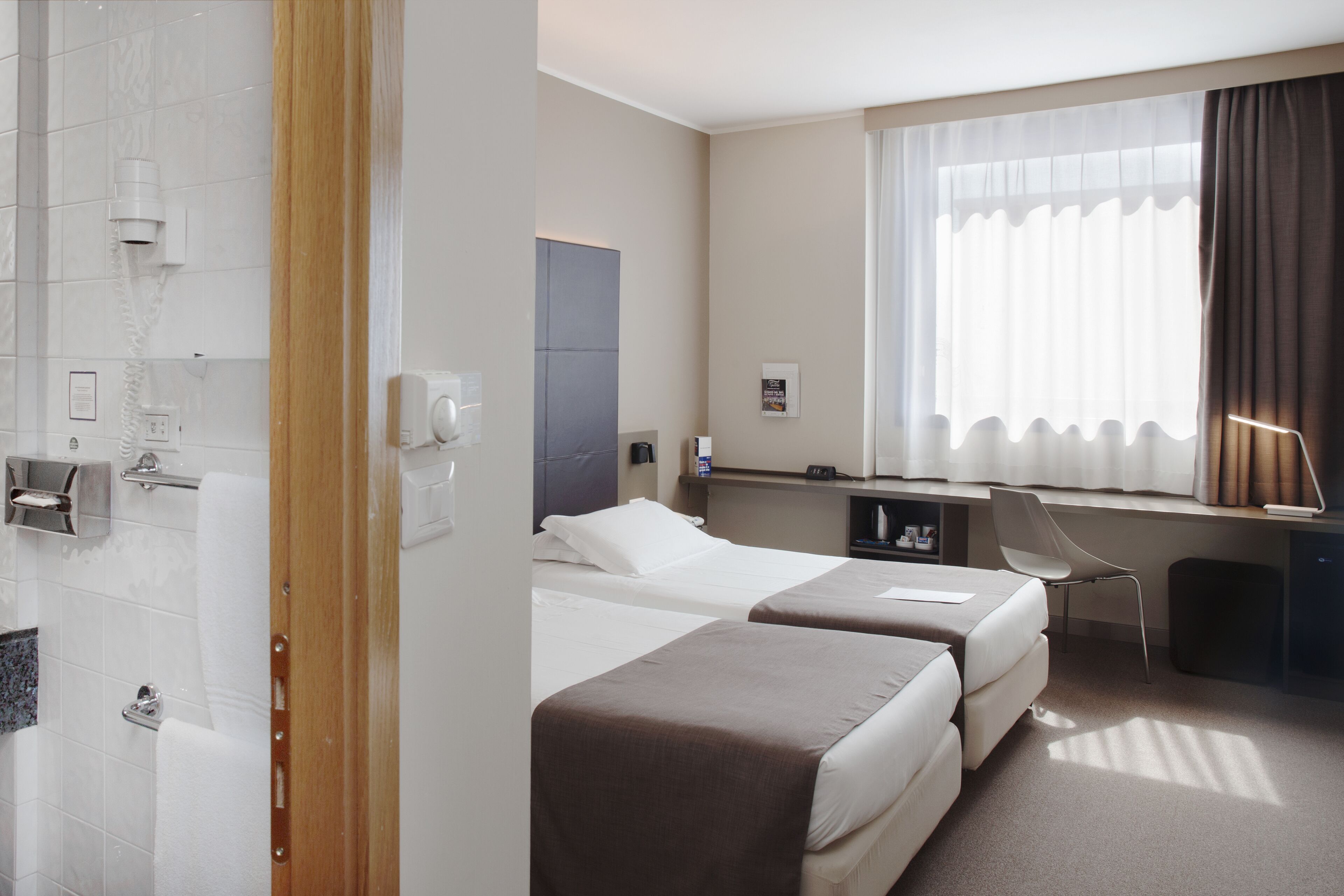 A hotel room with two beds, a desk, a chair, a shower, and a window with curtains.
