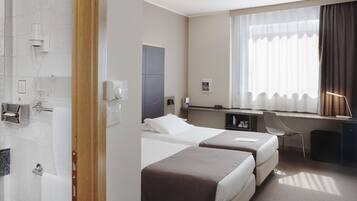 Habitación de hotel con dos camas, un escritorio, una silla, ducha y ventana con cortinas.