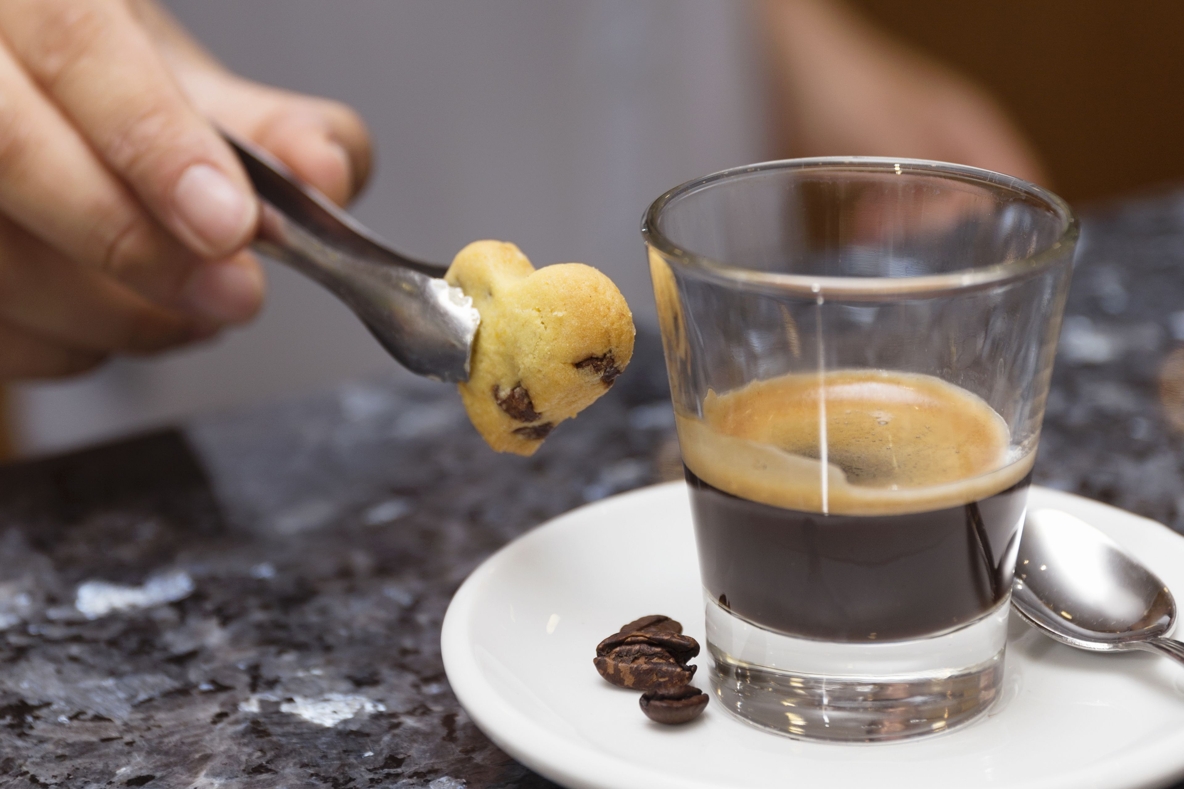 A spoonful of food being dipped into a glass of dark liquid, possibly coffee, on a white plate with a spoon beside it.