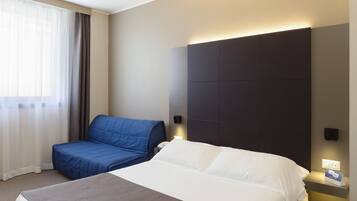 A hotel room with a large bed, a smaller bed, a bedside table with a lamp, a window with curtains, and a wall-mounted light.