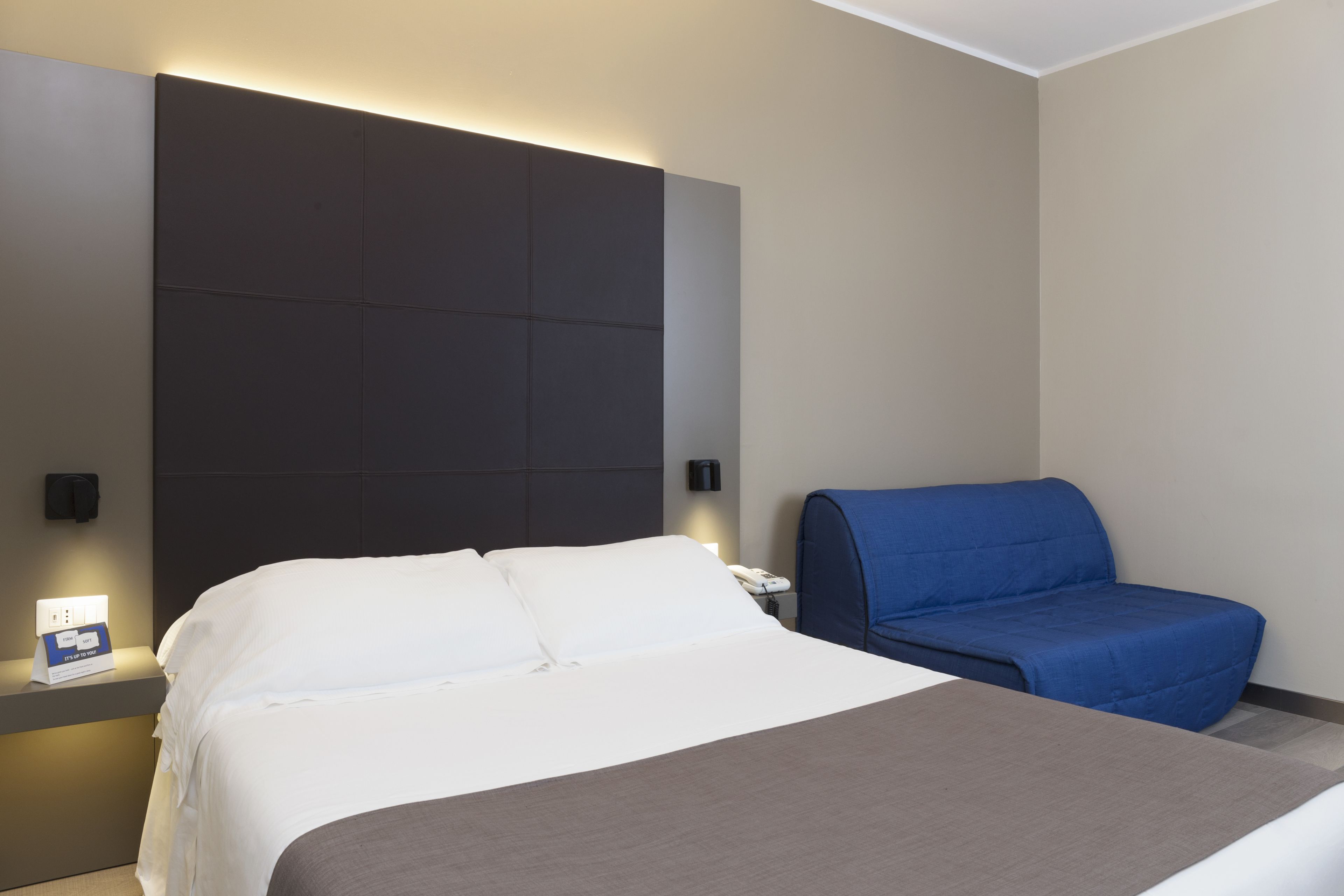 A modern hotel room with a large bed, a blue armchair, and a bedside table with a lamp and a phone.