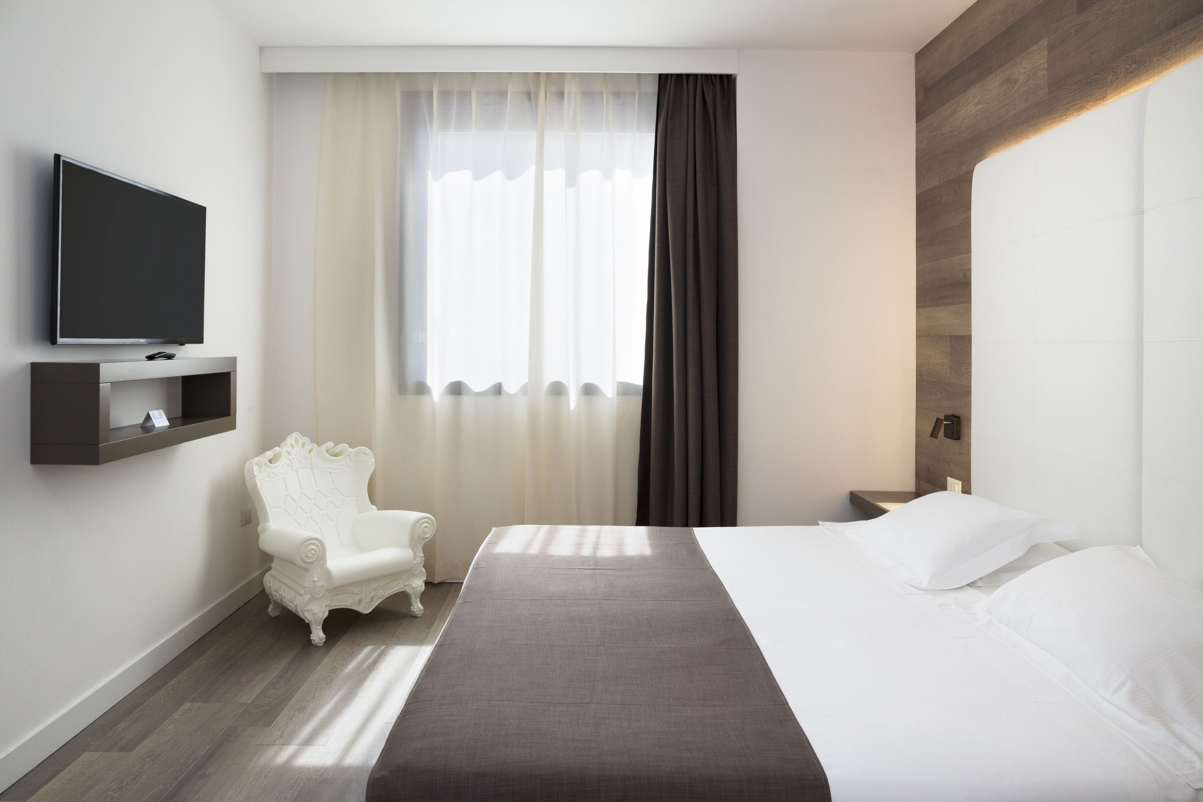 Una habitación de hotel con una cama, televisión montada en la pared, un sillón blanco y una ventana con cortinas.