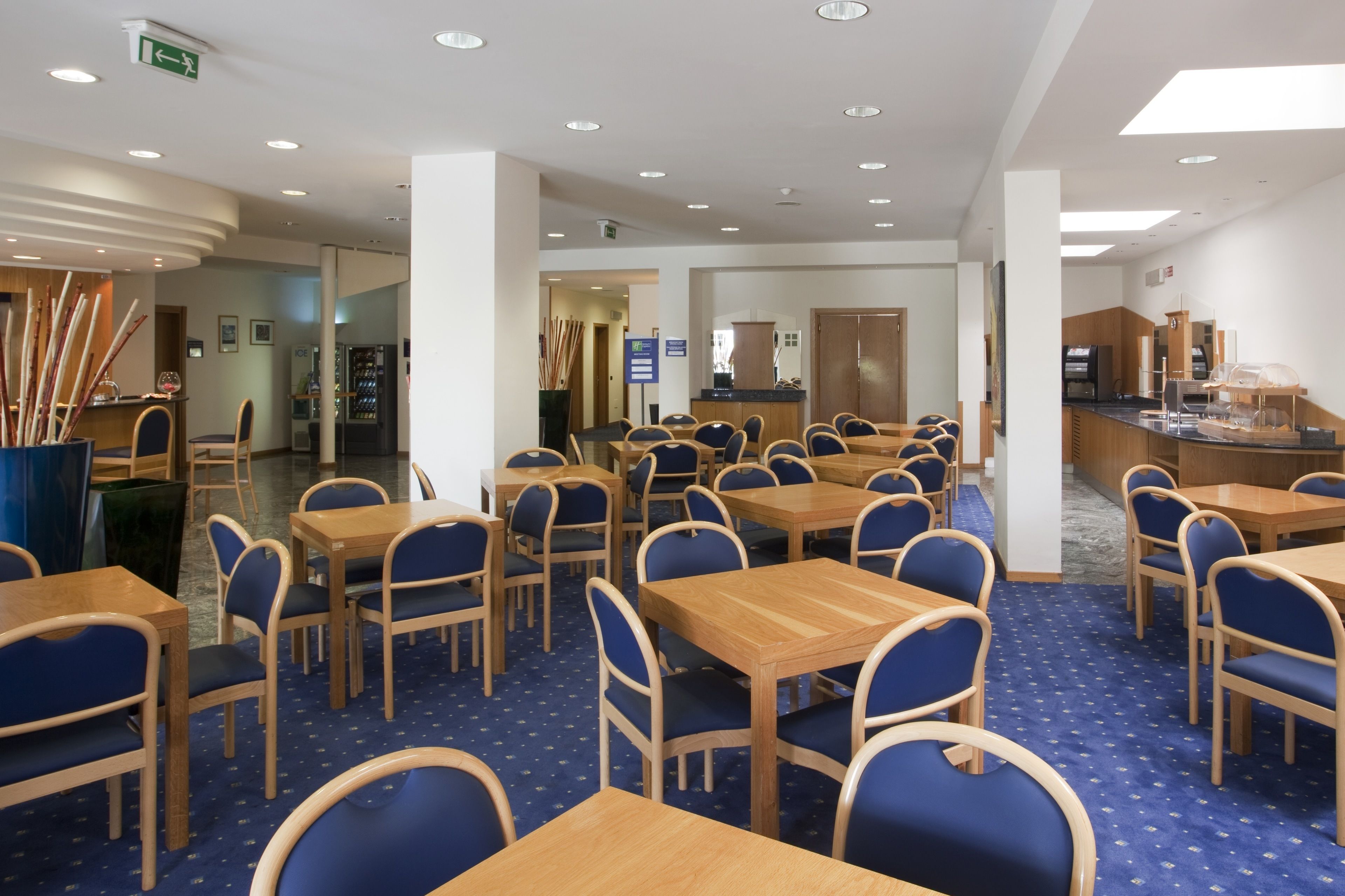 A hotel dining area with multiple tables and chairs, a reception desk, and a kitchen in the background.
