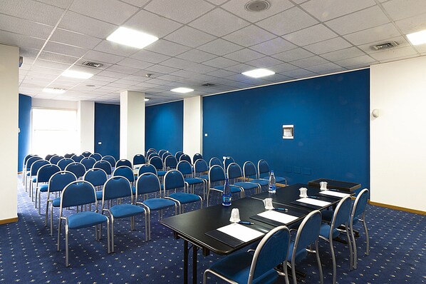 A conference room with rows of blue chairs, tables with water bottles, and a blue carpeted floor.