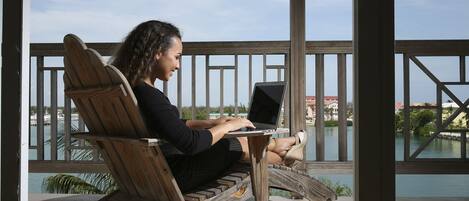 A person sitting on a wooden deck chair with a laptop, overlooking a body of water and buildings.