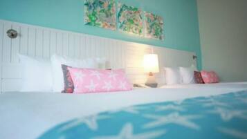 Quarto com cabeceira branca, cama com colcha estampada em azul e branco, e várias almofadas, incluindo uma rosa com padrão de estrela.