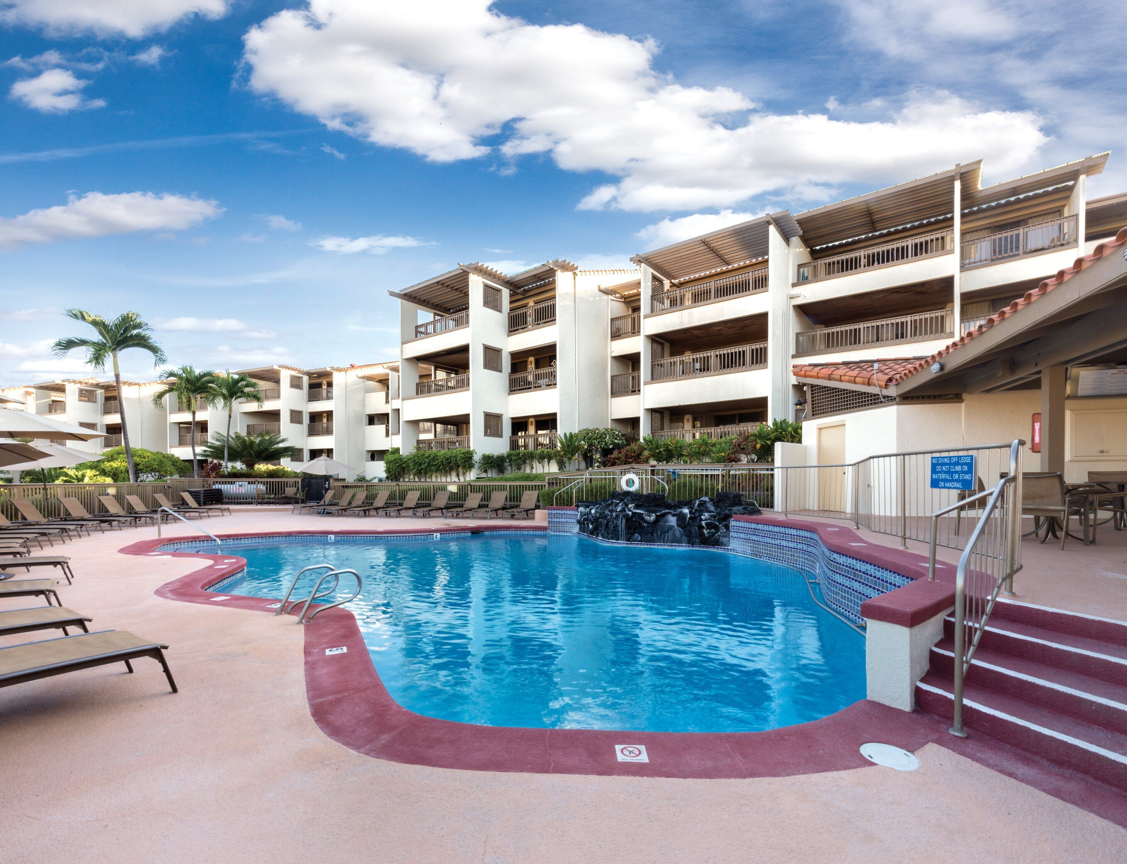 Un área de piscina con sillones de descanso, una piscina y un edificio de varios pisos con balcones.