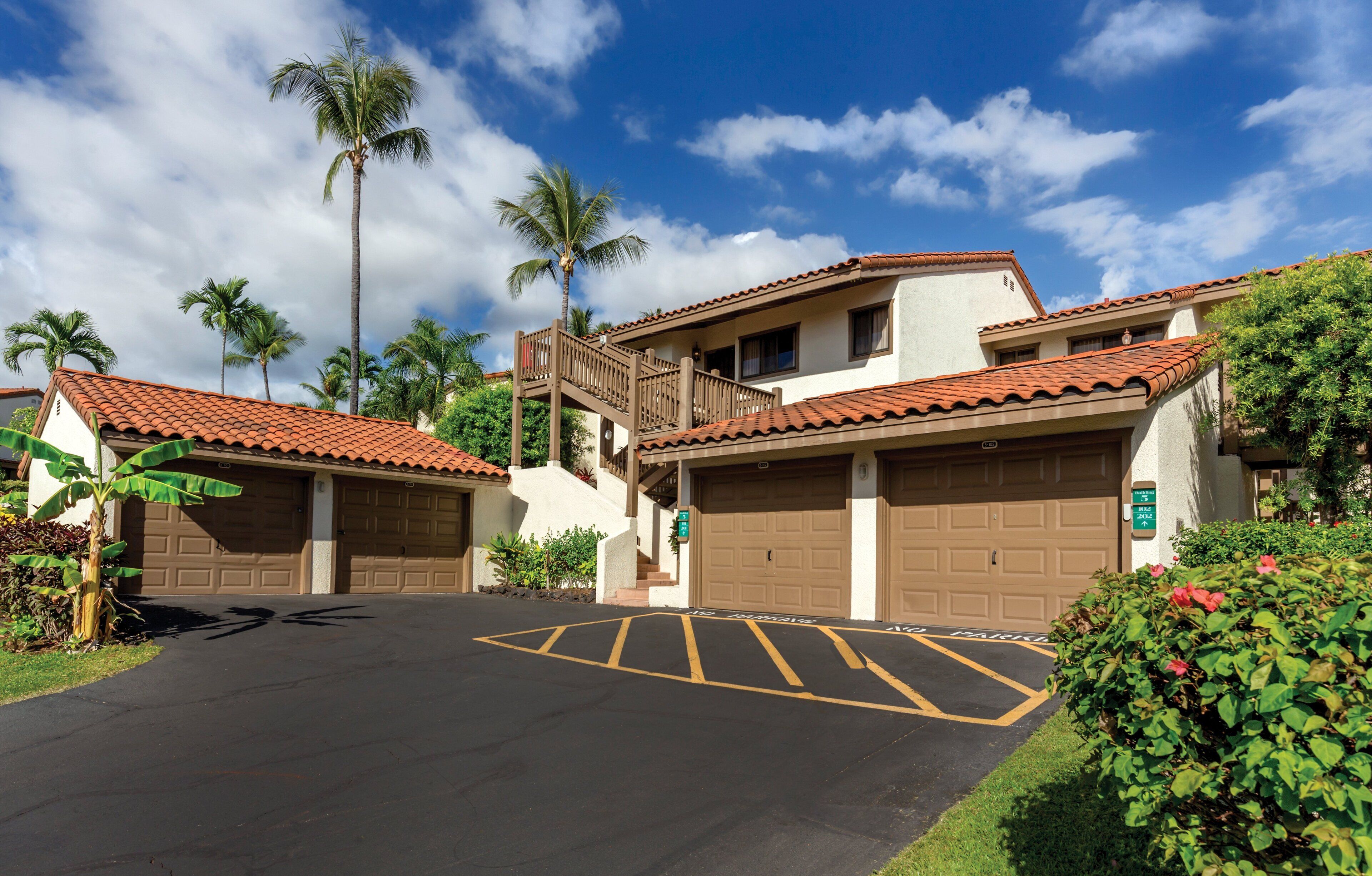 A residential area with two detached houses, each with a garage, surrounded by lush greenery and palm trees.