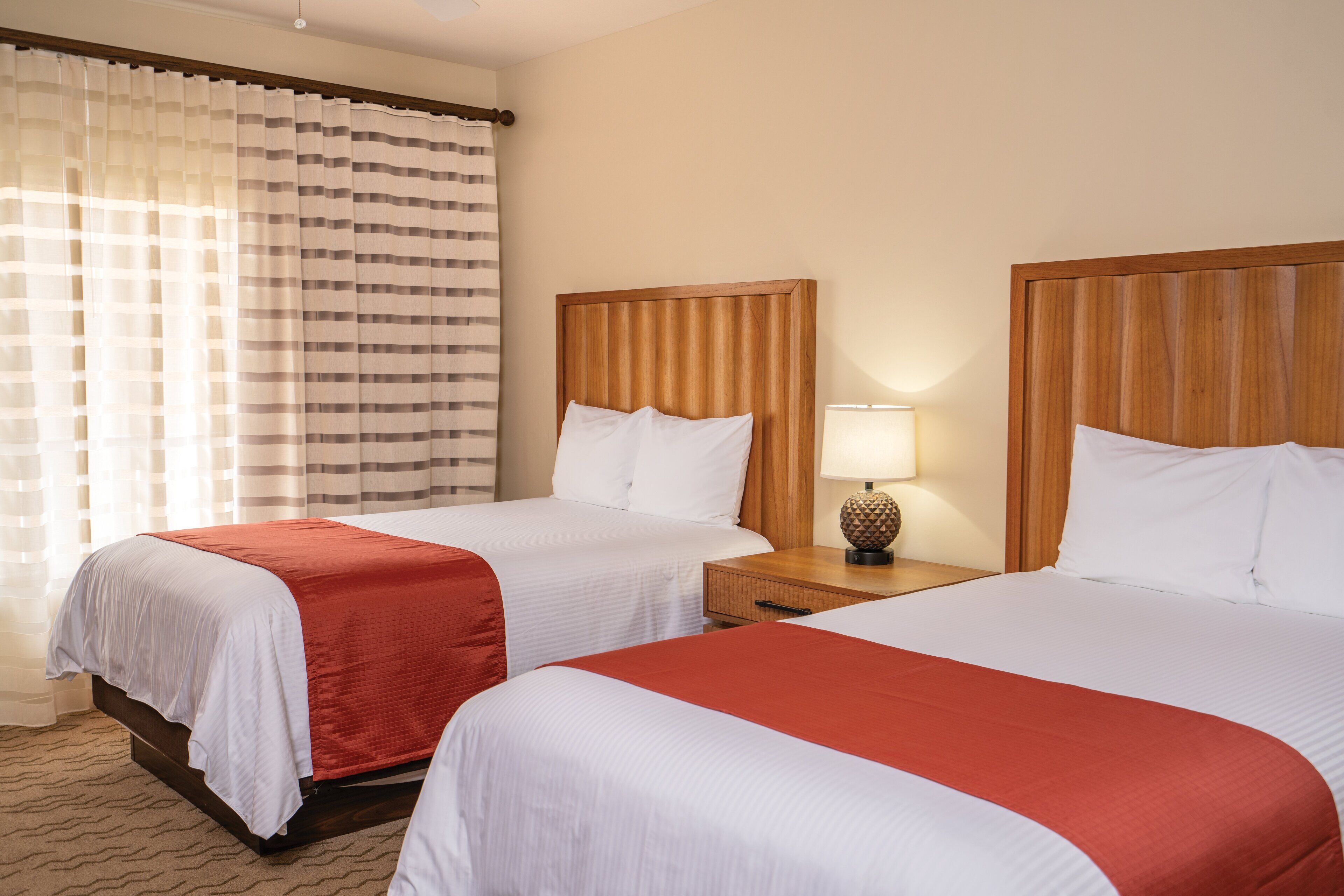 Une chambre d’hôtel avec deux lits, chacun doté d’une tête de lit en bois, de draps blancs et d’un élément décoratif rouge. Une table de chevet avec une lampe se trouve entre les deux lits.
