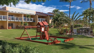 A playground with a slide and climbing structure, surrounded by a well-maintained lawn and palm trees, in front of a yellow building with multiple balconies.