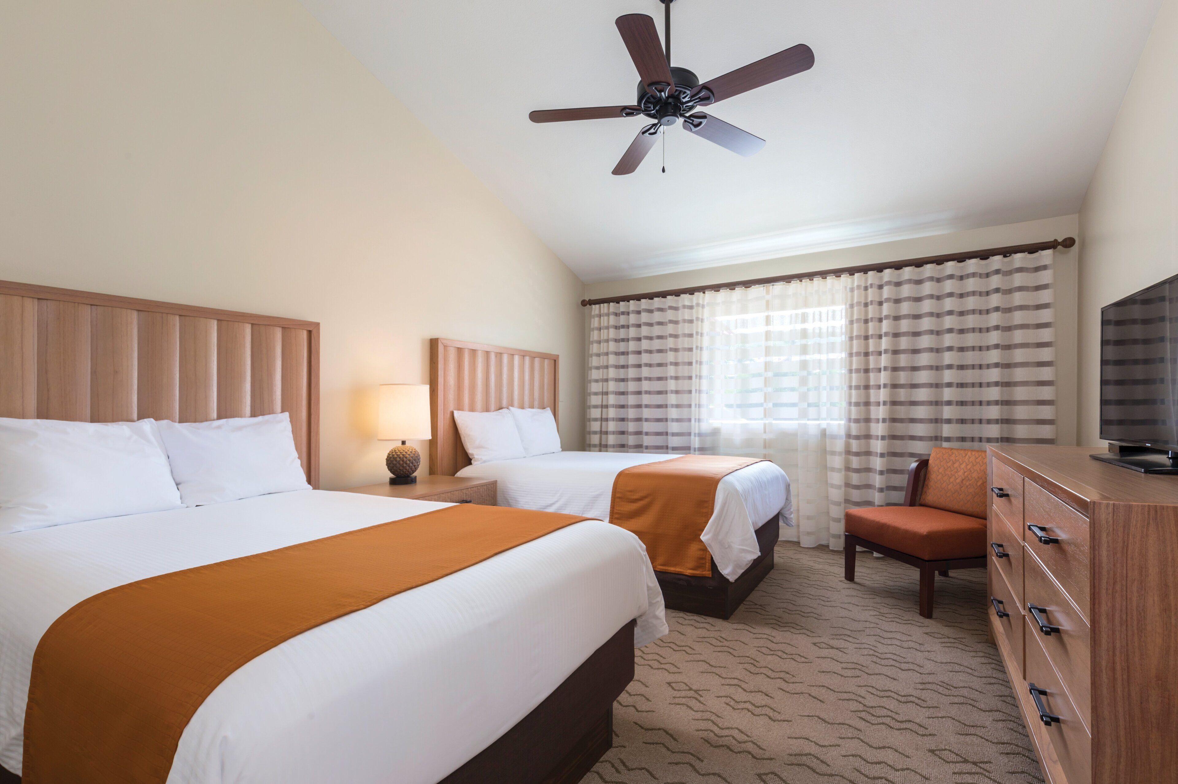 A hotel room with two beds, a ceiling fan, a TV, a chair, and a dresser.