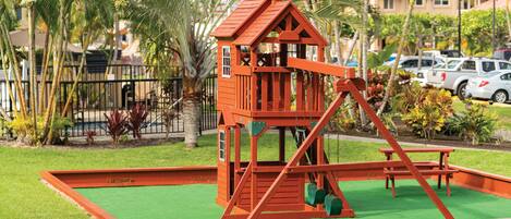 A red wooden play structure with a slide and swings, surrounded by green artificial grass and palm trees.
