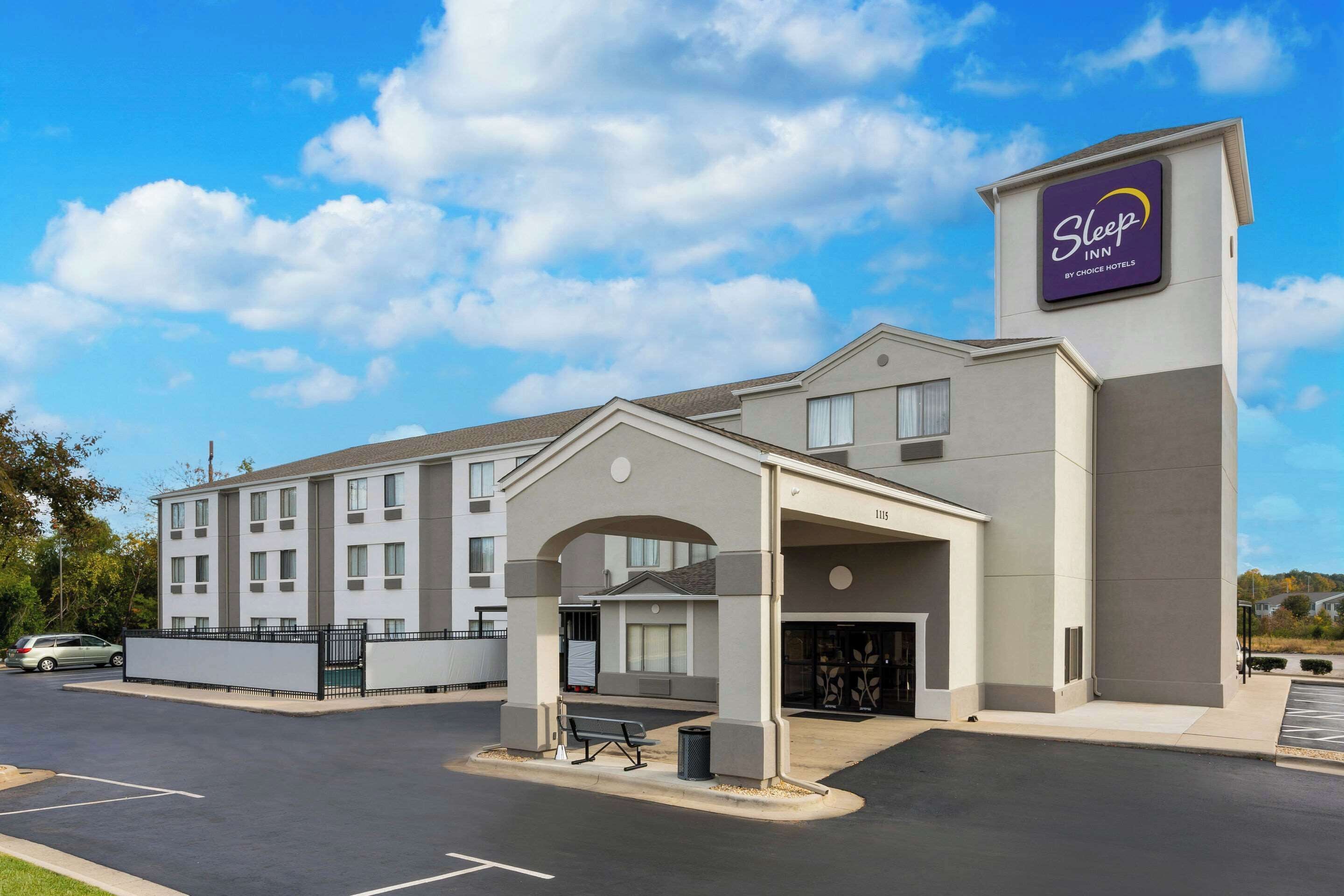 A Sleep Inn by Choice Hotels building with a covered entrance, a parking lot, and a clear sky.