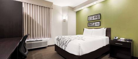 A hotel room with a bed, a desk, a chair, a nightstand, and a wall-mounted air conditioner.