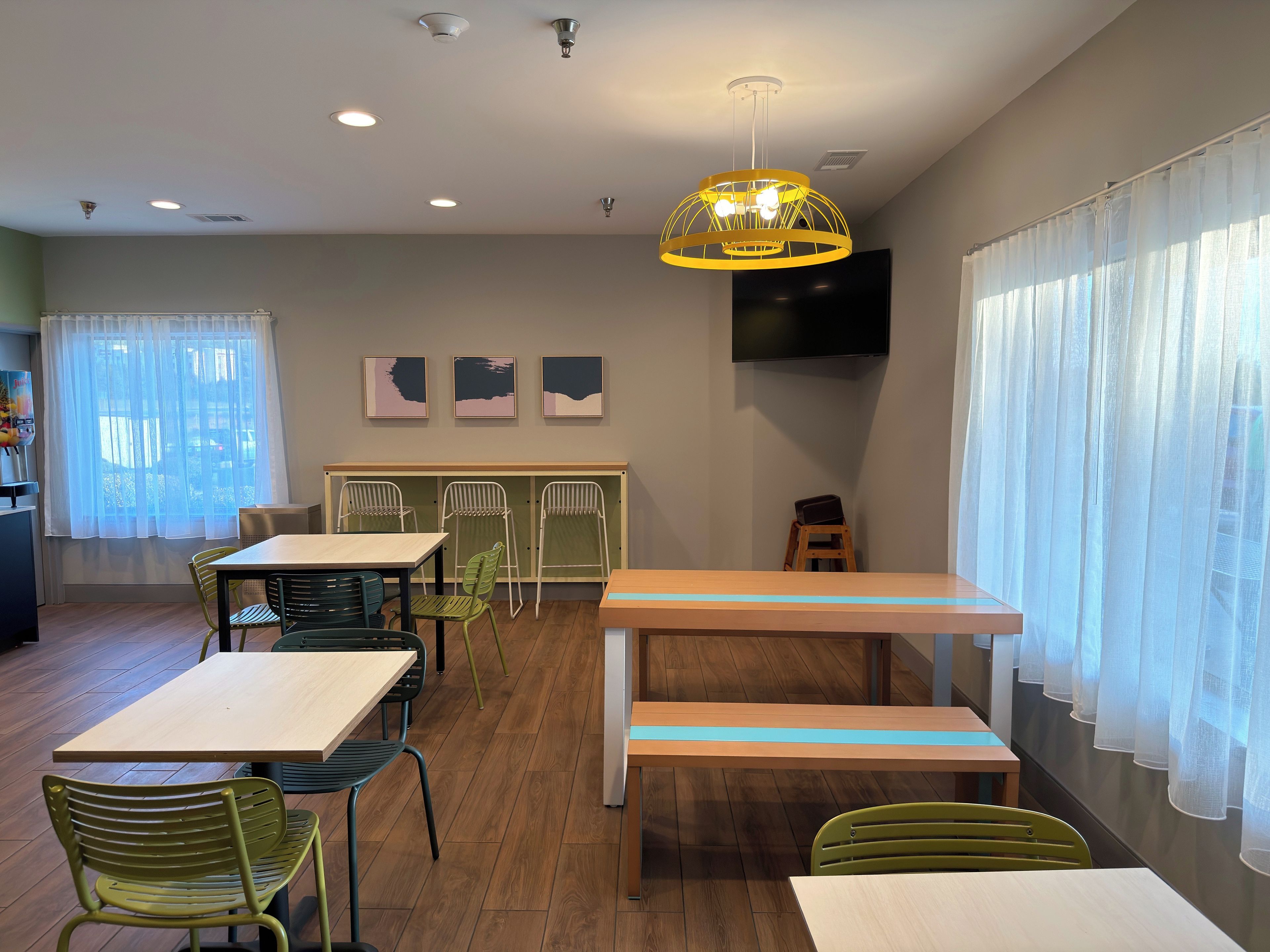 A modern dining area with tables, chairs, a bench, a ceiling light fixture, and a flat-screen TV mounted on the wall.