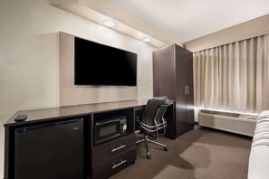 A hotel room with a flat-screen TV mounted on the wall, a desk with a microwave, a chair, a wardrobe, and a large window with curtains.