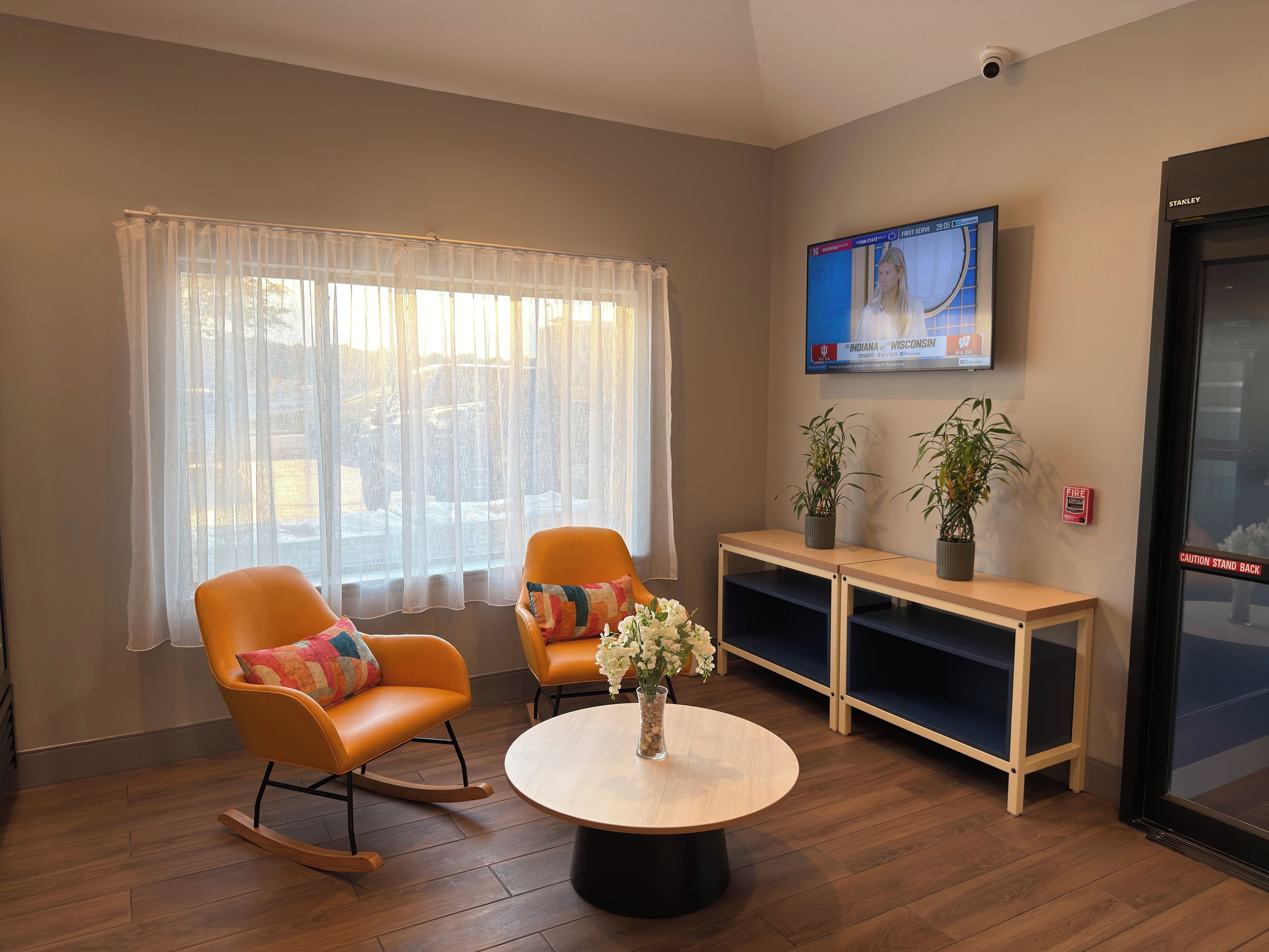 Een kamer met twee schommelstoelen, een ronde tafel met een vaas, een televisie aan de muur en een brandalarm.