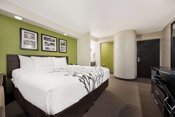 A hotel room with a large bed, a desk, and a chair. There are framed pictures on the wall and a door to the right.