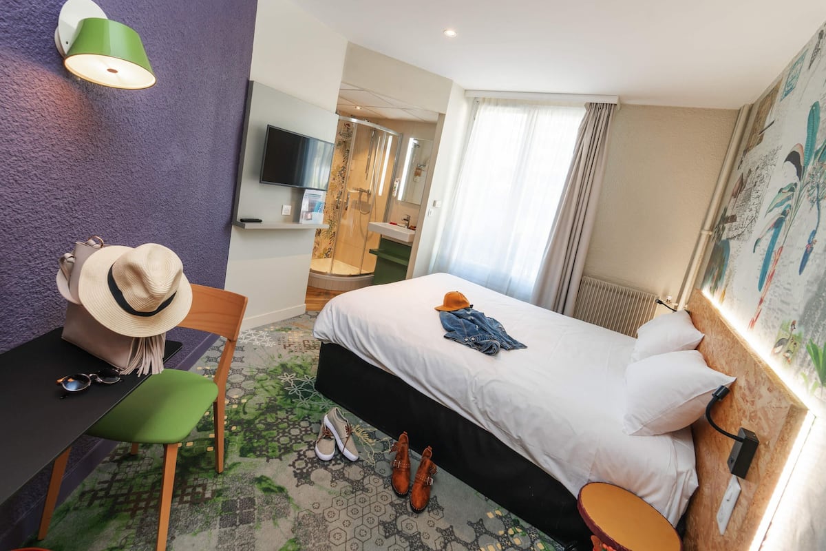 Hotelkamer met een bed, bureau met hoed, stoel, televisie, douche en raam met gordijnen.