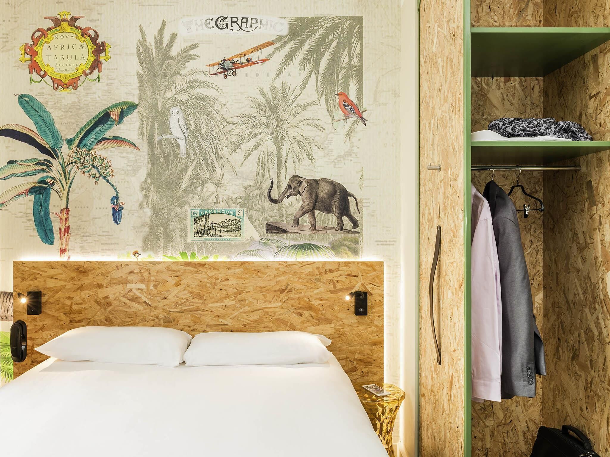 A room with a bed, a wooden headboard, a wall with a map and illustrations, a green shelf with clothes, and a small table with a lamp.