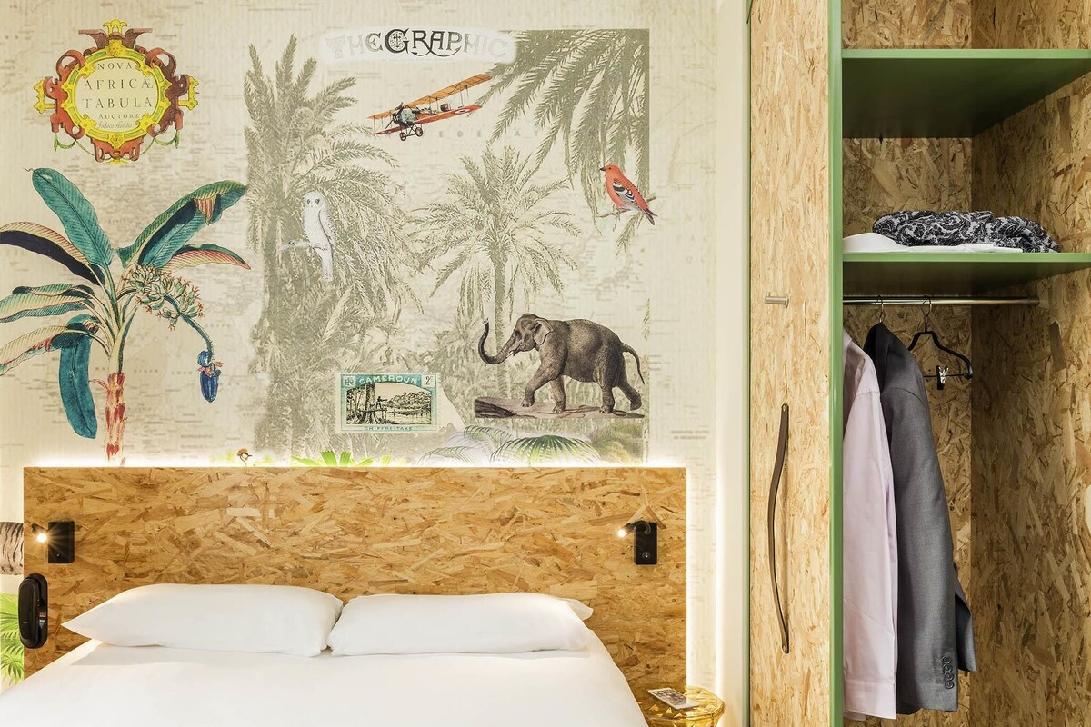 Une pièce comprenant un lit, une tête de lit en bois, un mur orné d’une carte et d’illustrations, une étagère verte avec des vêtements et une petite table avec une lampe.