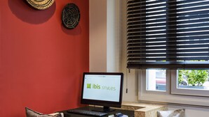 Un comptoir d’accueil avec un ordinateur affichant le logo « ibis styles », une chaise en bois, une petite table et une fenêtre avec des stores.
