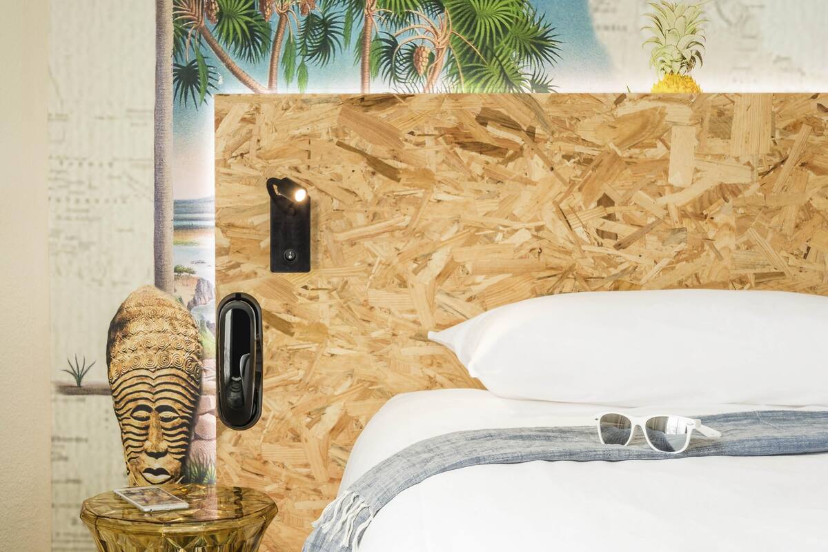 Een kamer met een houten wand, een bed met witte beddengoed, een klein tafeltje met een decoratief object, en een wandlamp.