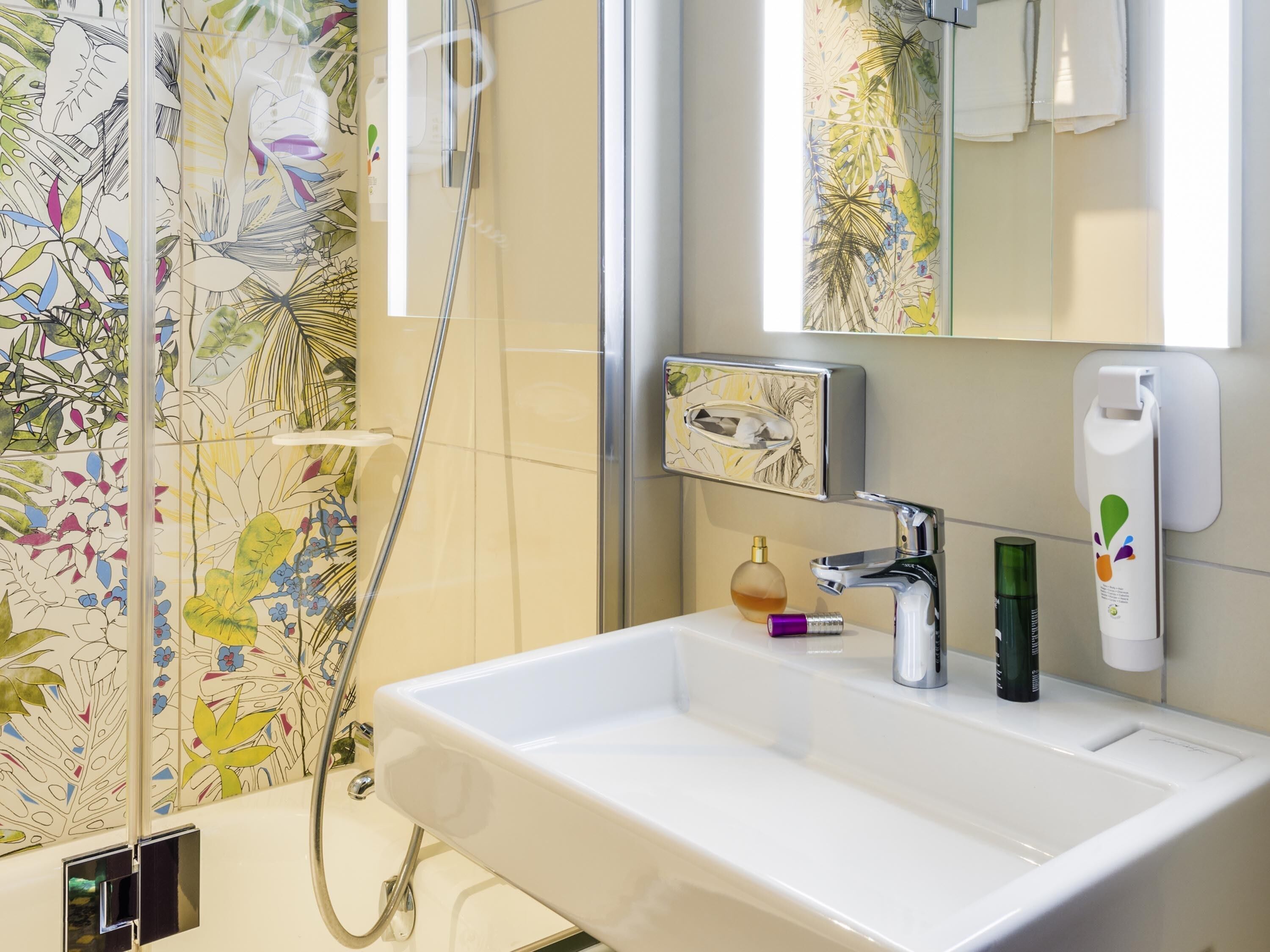 Ein Badezimmer mit weißem Waschbecken, einem Spiegel, einer Dusche und einer Wand mit floralem Fliesendesign.