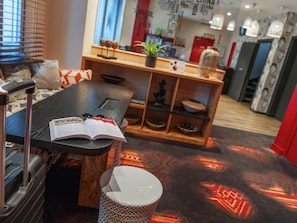 Quarto de hotel com uma cama, uma secretária com um laptop e um livro, uma mala, um banquinho pequeno, uma estante com objetos decorativos e um tapete estampado.