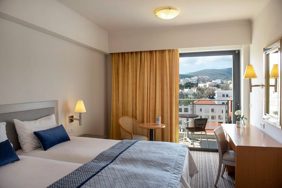 A hotel room with two beds, a desk, a chair, a small table, and a view of the city through a large window.