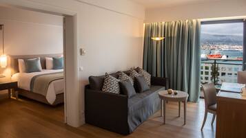 A modern hotel room with a bed, a sofa, a small table, and a view of a cityscape through a large window.