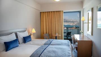 A hotel room with two beds, a desk, a chair, a small table, and a view of a coastal town.