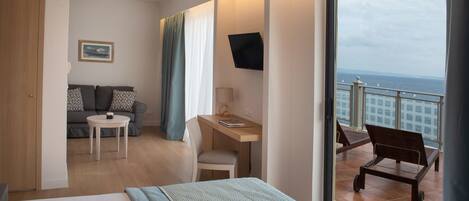 A hotel room with a bed, a desk, a chair, a sofa, and a balcony with a view of the sea.