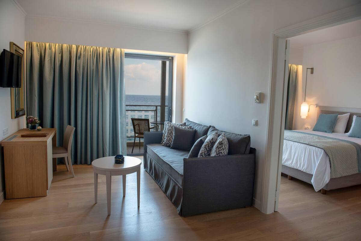 A modern hotel room with a sofa, a small round table, a bed with a view of the sea, and a balcony with curtains.