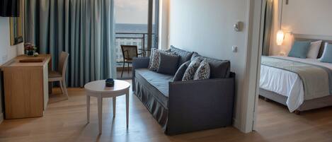 Quarto de hotel moderno com sofá, uma pequena mesa redonda, cama com vista para o mar e varanda com cortinas.