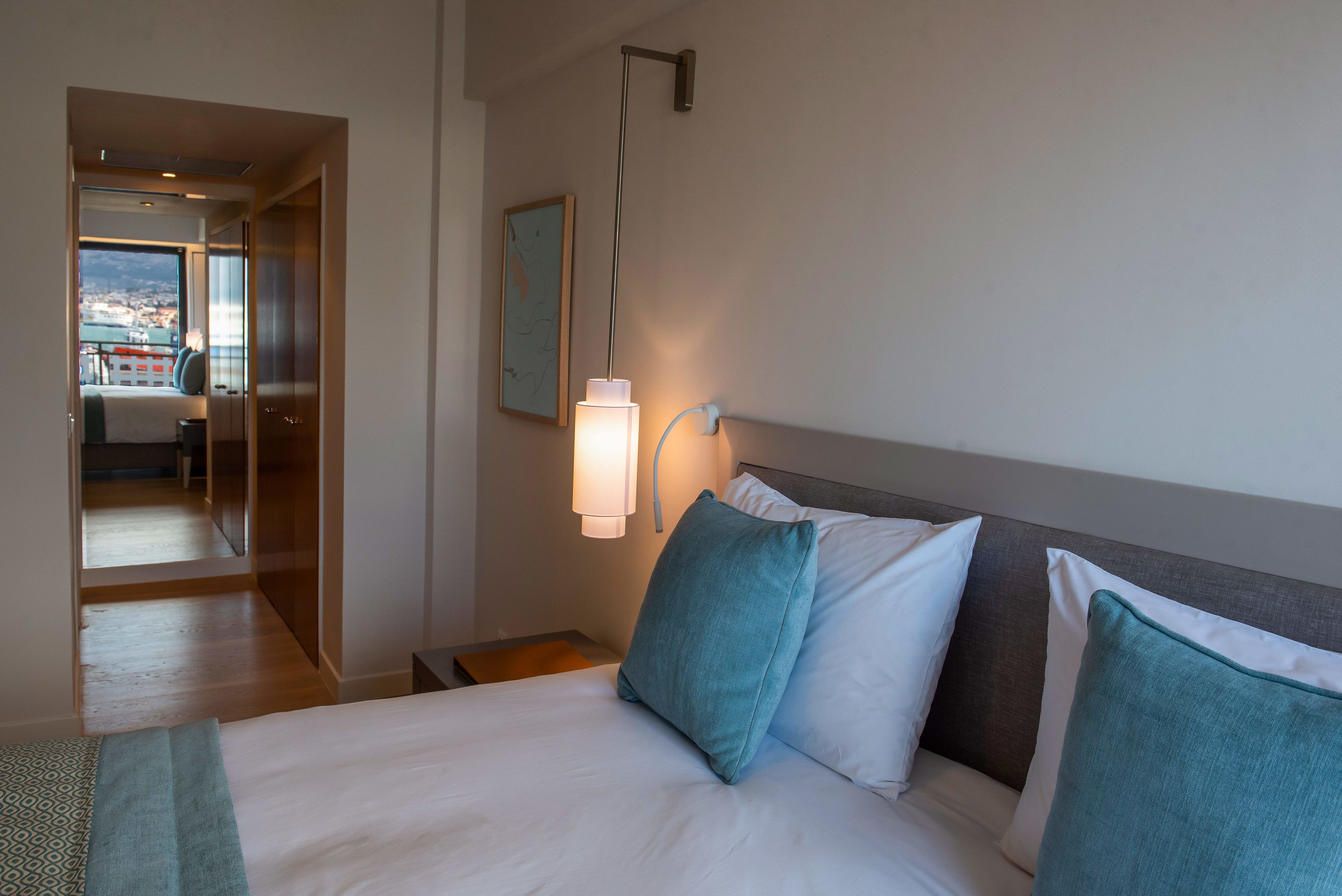Ein Hotelzimmer mit Bett, Nachttisch, Lampe und Blick auf die Stadt durch ein großes Fenster.