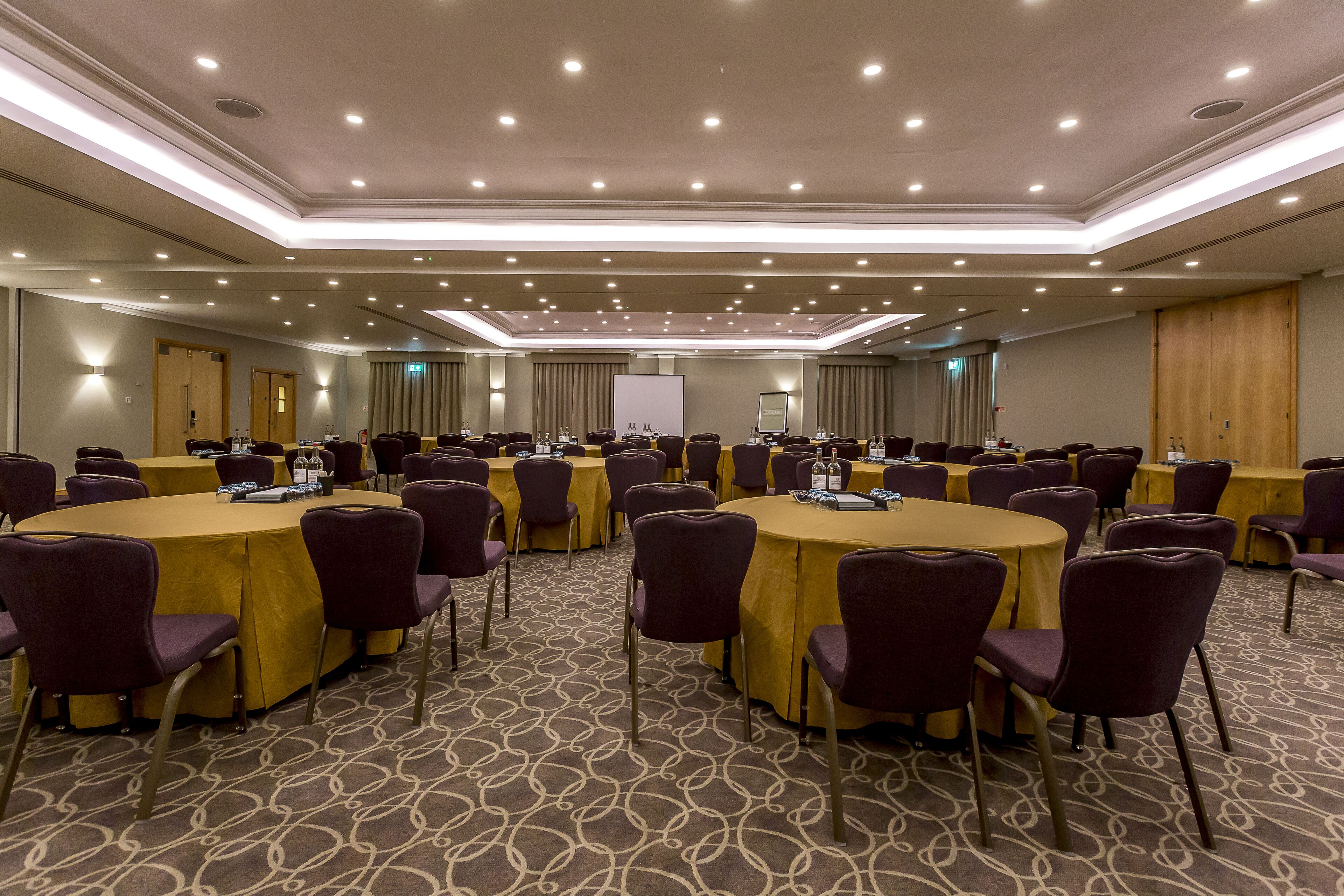 Une salle de conférence dotée de plusieurs tables rondes, chacune étant entourée de chaises, et équipée de bouteilles d’eau et de porte-noms.