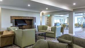 A spacious lobby with a fireplace, comfortable seating, and a dining area in the background. - Hellidon Lakes Golf & Spa Hotel (Daventry)