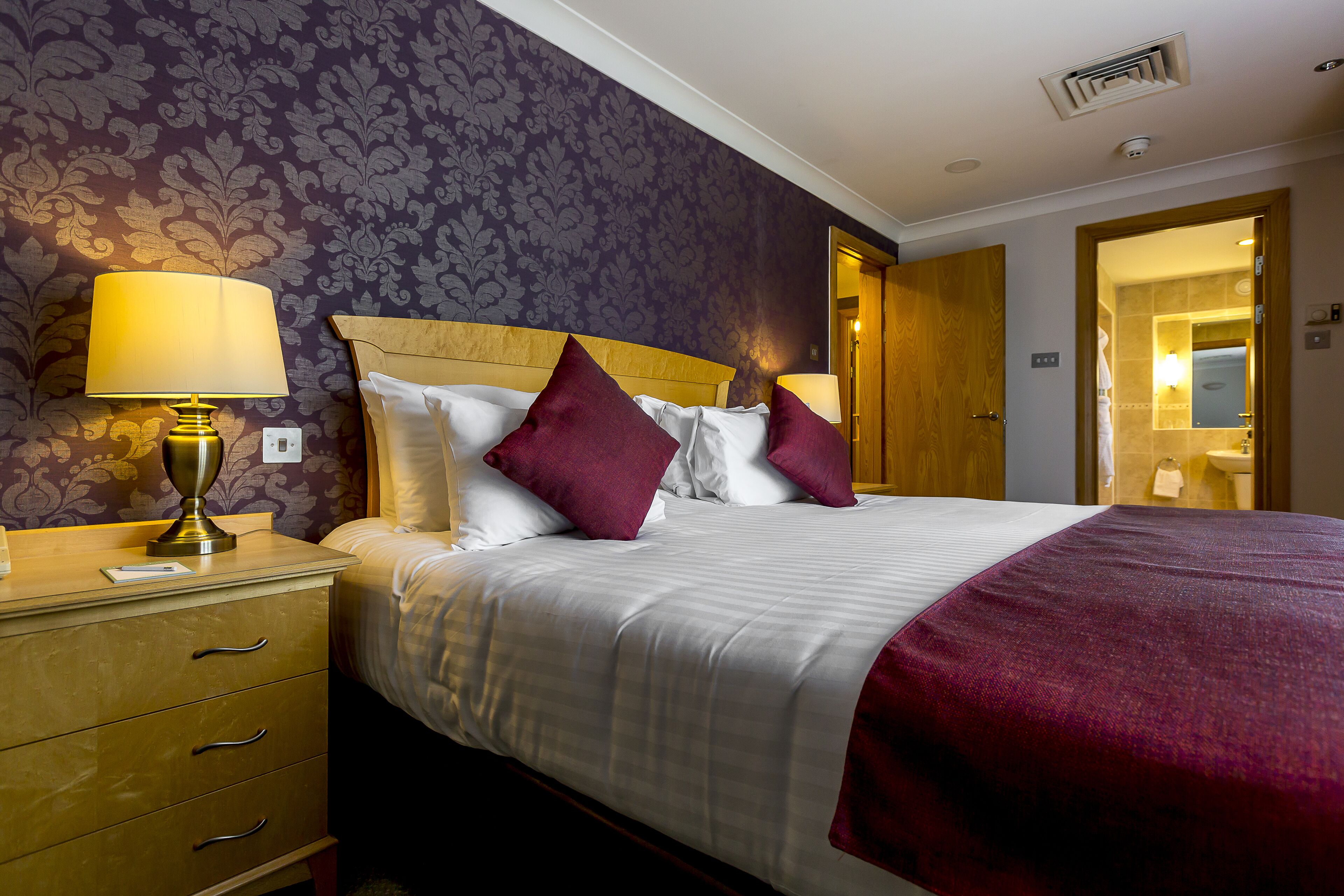 A hotel room with a bed, a nightstand with a lamp, a wooden headboard, and a door leading to a bathroom.