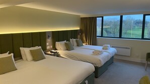 A hotel room with two beds, a nightstand, a telephone, and a view of a green landscape through large windows. - Hellidon Lakes Golf & Spa Hotel (Daventry)