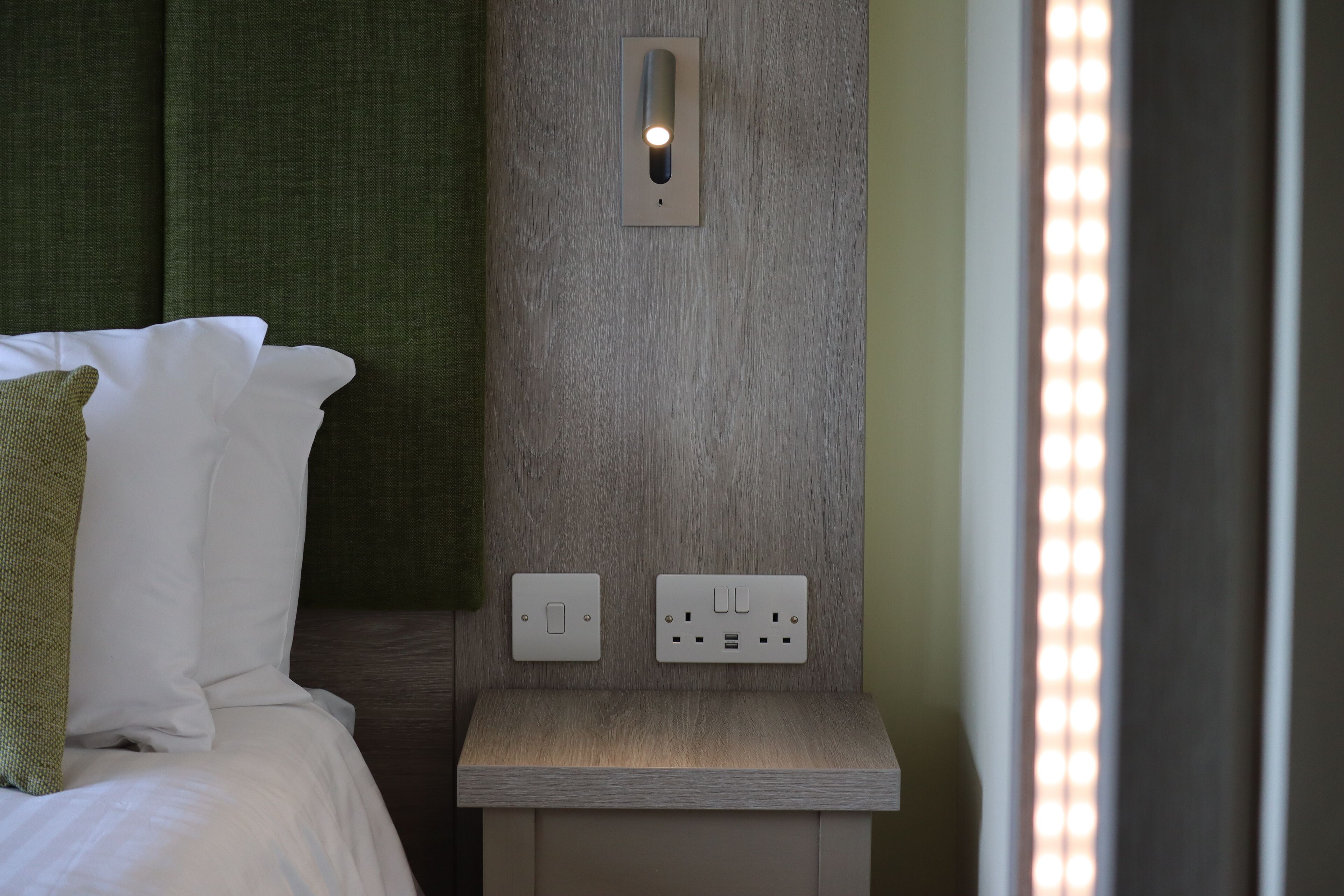 A neatly made bed with white linens and a green pillow, a wall-mounted light fixture, and a power outlet on a bedside table.