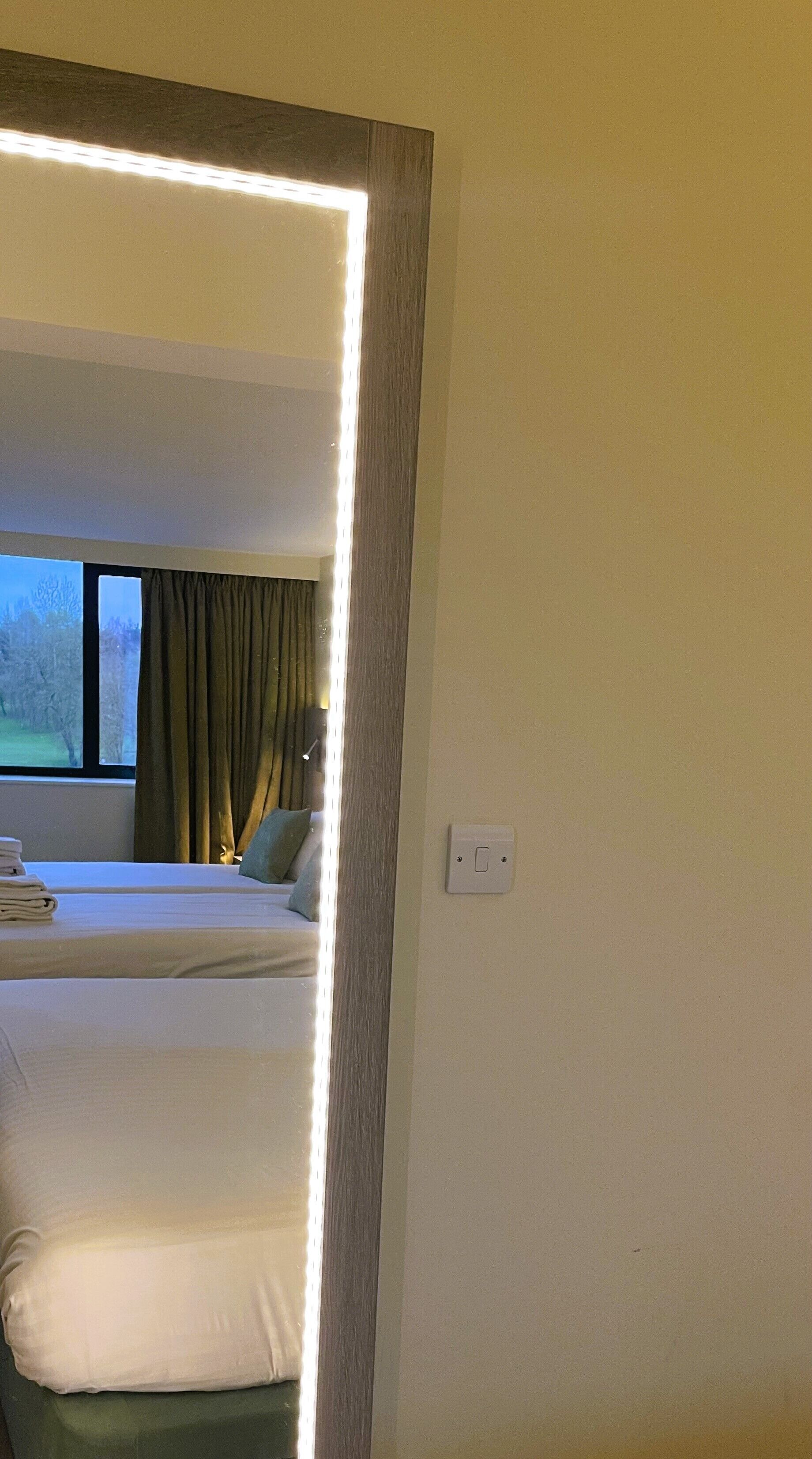 A hotel room with a large mirror reflecting a bed, a window with curtains, and a view of trees outside.