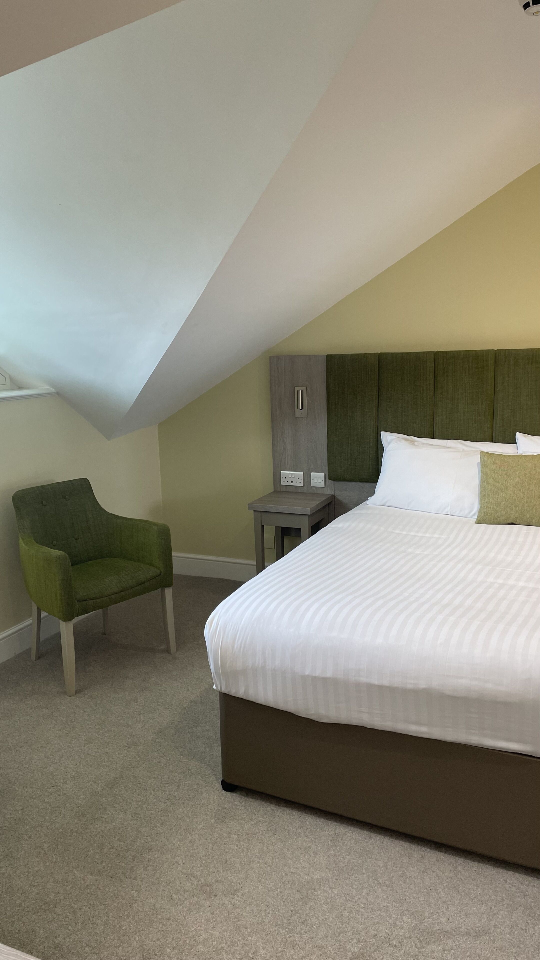A hotel room with a large bed, a green armchair, a nightstand, and a wardrobe.