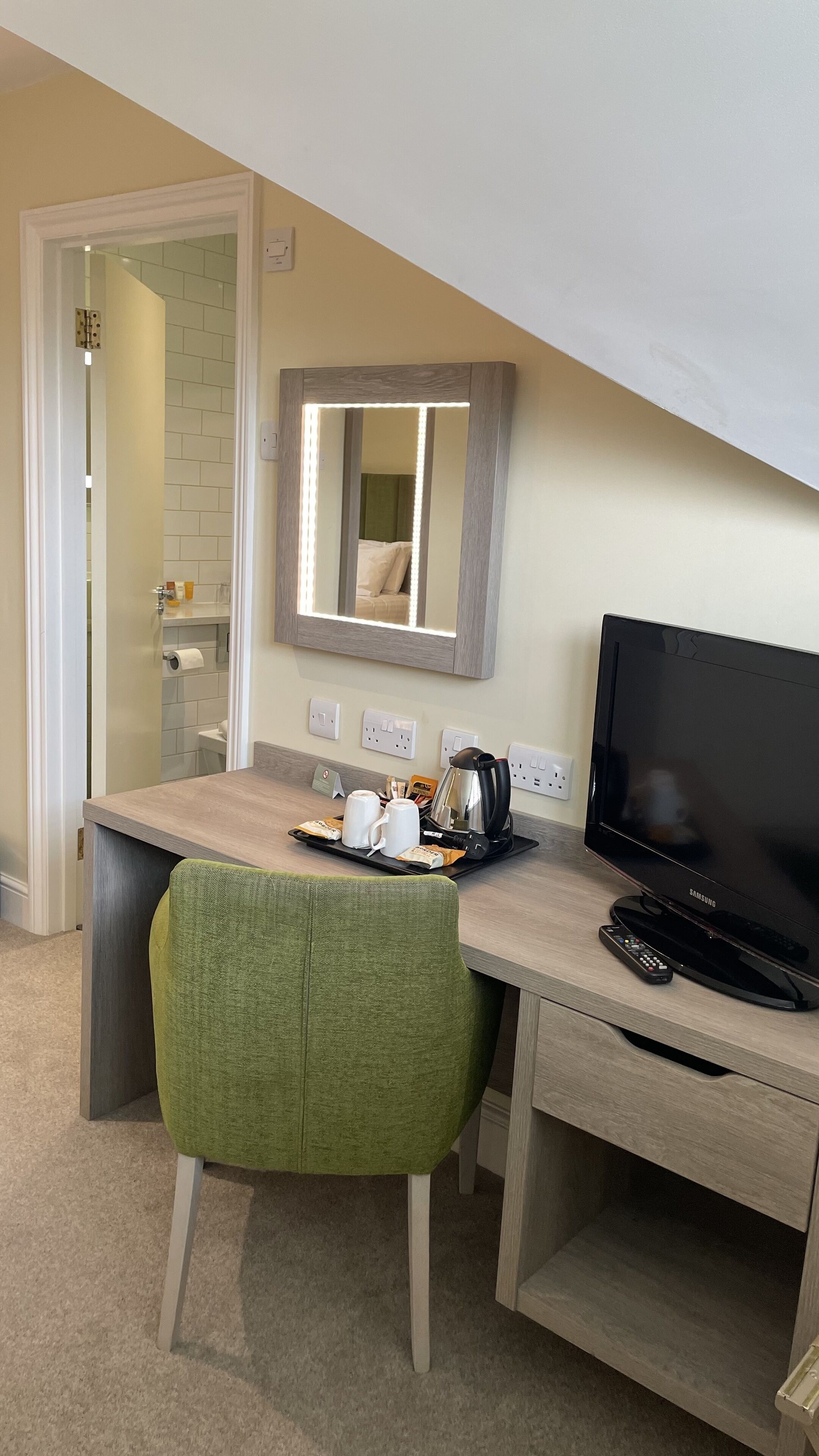 A hotel room with a desk, a green chair, a television, a coffee maker, and a tray with cups and a remote control.